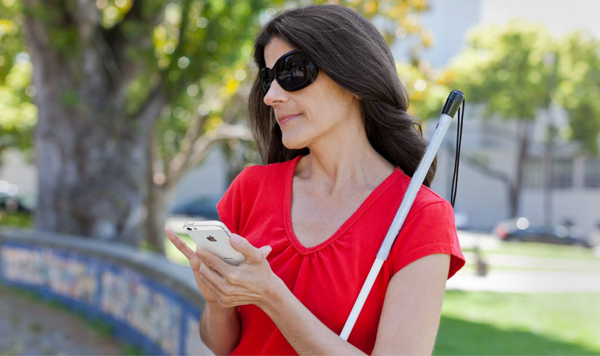 Acessibilidade: mulher cega, usando óculos escuros e abraçada a uma bengala utiliza seu smartphone em meio a natureza.