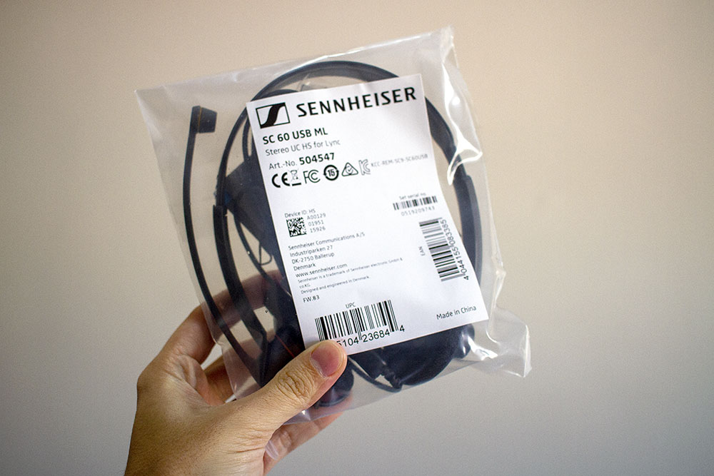 Mão segurando a embalagem do Headset Sennheiser: um plástico simples envolvendo todos os itens e a frente, um adesivo branco com escritos simples, em preto, com características do produto