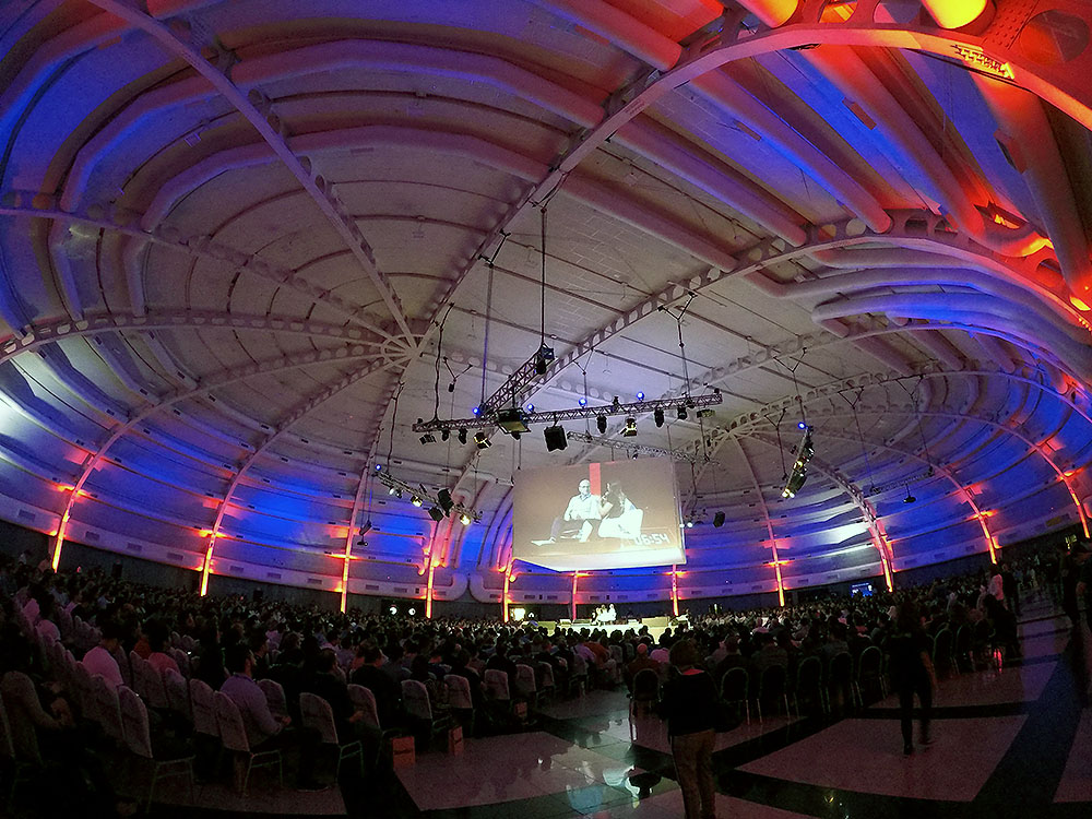 Estrutura de ferro grande com iluminação colorida, vermelha e azul, um enorme telão sobre os convidados e mais de 2.000 participantes em volta
